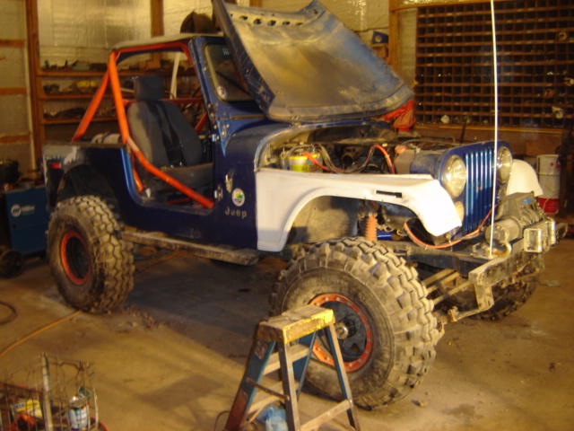 Jeep CJ with hood open, blue body, orange roll cage, and large off-road tires. It's in a garage with tools visible