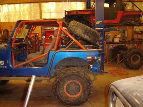 1992 Ford Bronco - A blue Jeep with an orange roll cage and a spare tire mounted on the back is in a garage, with another vehicle on a lift above it.