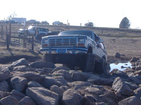 1986 Ford Bronco - A Ford Bronco drives over rocks with other vehicles in the background.