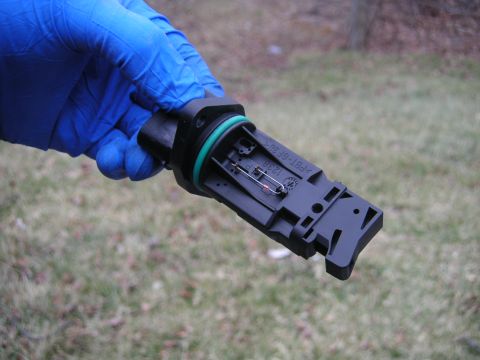 2002 BMW M3 - A black plastic mass air flow sensor with a green O-ring is held by a gloved hand against a blurred outdoor background.
