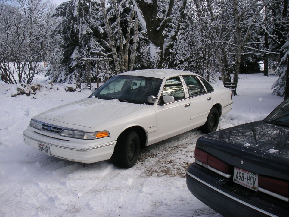 White Ford Crown Victoria on steel wheels parked in snow beside a dark blue sedan