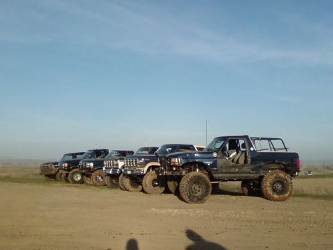 1995 Ford Bronco - A convoy of modified Ford Broncos and trucks are lined up on a dirt lot under a clear blue sky.