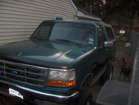 1996 Ford Bronco - The front end of a green Ford Bronco is visible, parked in a residential area with a fence and dog in the background.
