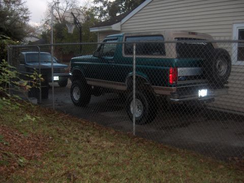 1996 Ford Bronco - A green Ford Bronco with a tan removable top is parked behind a fence, with another vehicle visible in the background.