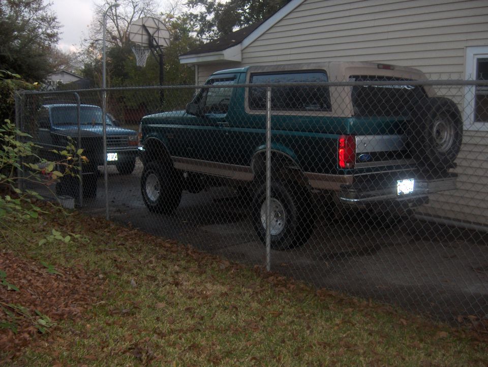 Green Ford Bronco with tan top and spare tire on rear, parked behind a chain-link fence