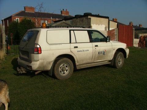 2006 Mitsubishi L200 - A white panel van is parked on a grassy area in front of some houses. The van has company markings on its side.
