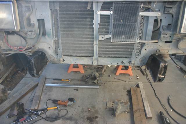 Front end of a vehicle disassembled, with radiator and AC condenser exposed. Tools and jack stands are on the ground