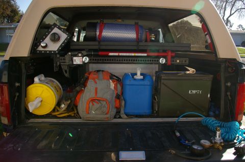 1986 Ford Bronco - The rear of a truck is loaded with various equipment, including a blue water jug, a backpack, and a green ammo can labeled 'EMPTY'.