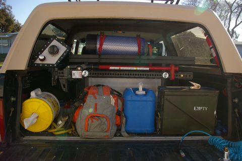 1986 Ford Bronco - The rear of a truck bed is organized with off-road equipment, including a Hi-Lift jack, water container, and storage boxes.