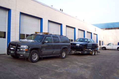 2001 Ford Crown Victoria - A dark blue 2001 Ford Crown Victoria is transported on a trailer, with a dark SUV in front and a white pickup truck in the background.
