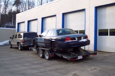 2001 Ford Crown Victoria - A blue and gray 2001 Ford Crown Victoria is loaded onto a trailer, with a gray SUV visible in front.