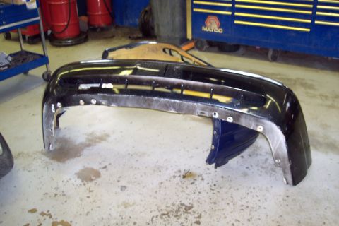 2001 Ford Crown Victoria - A detached black bumper lies on the floor of a workshop, with tools and equipment in the background.