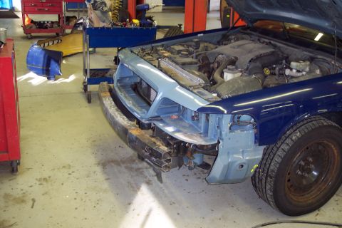 2001 Ford Crown Victoria - Front end of a blue 2001 Ford Crown Victoria undergoing repairs in a workshop, showing damage and exposed engine components.