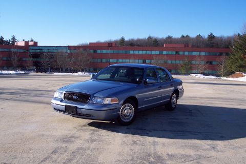 2001 Ford Crown Victoria - A blue 2001 Ford Crown Victoria is parked in an empty lot on a sunny day, with a large red brick building behind it.