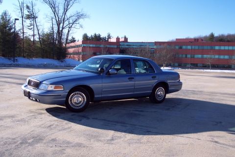 2001 Ford Crown Victoria - This blue 2001 Ford Crown Victoria sedan is seen from a three-quarter front view in a parking lot with a modern building in the background.