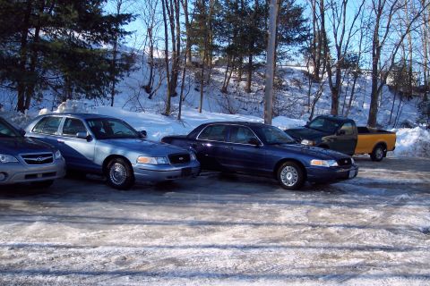 2001 Ford Crown Victoria - Two blue sedans and a pickup truck are parked in a snowy lot, with the front sedan appearing to be a 2001 Ford Crown Victoria.