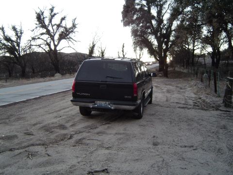 1989 Ford Bronco - A black GMC Yukon SUV is parked on a dirt road next to a paved road, with trees in the background.