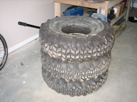 1996 Ford Bronco - Three chunky off-road tires are stacked in a garage, with a bicycle wheel peeking into the frame on the left.