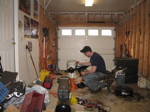 1995 Ford Bronco - A man works on an engine component inside a cluttered garage with a large overhead door.
