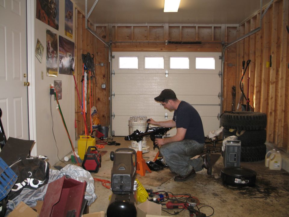Man in garage working on engine part, with tools and tires visible