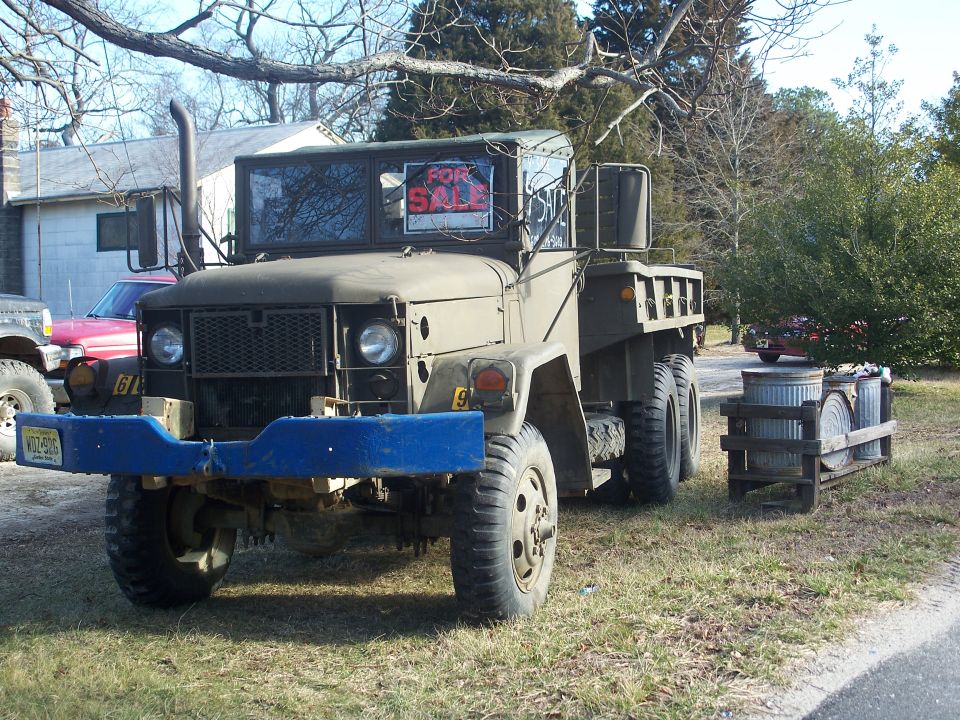 Military truck with blue bumper and 'For Sale' sign in window. Another vehicle is partially visible to the left