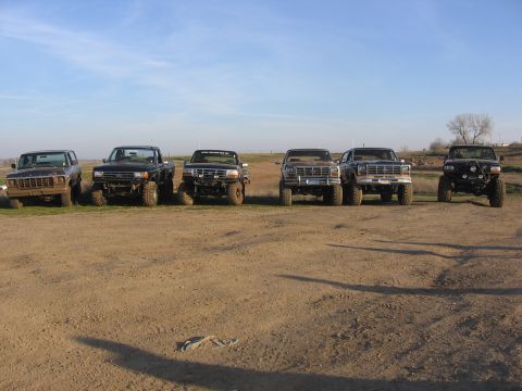 1989 Ford Bronco - A group of lifted trucks, likely from the late 1980s, are lined up in a dirt field under a clear blue sky.