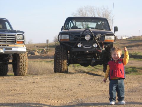 1989 Ford Bronco - Two lifted Ford trucks are parked on a dirt road with a young child posing in front of one.
