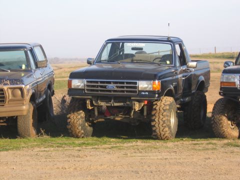 1989 Ford Bronco - Three lifted trucks are parked on a dirt lot with grassy areas under a clear sky.