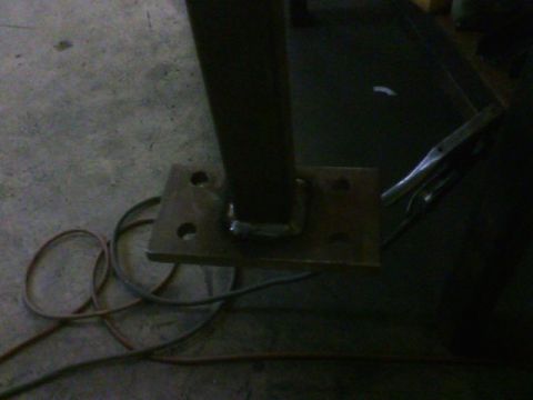 1990 Ford Bronco - A close-up view of a metal bracket with bolt holes, attached to a vertical post, with welding cables on the floor.
