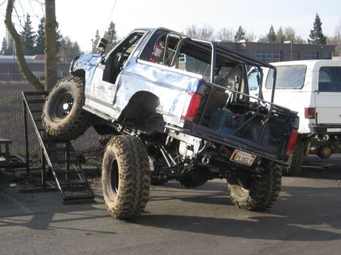 1994 Ford Bronco - Modified SUV with large tires on a ramp, showing custom roll cage and rear end. Another vehicle is parked behind.