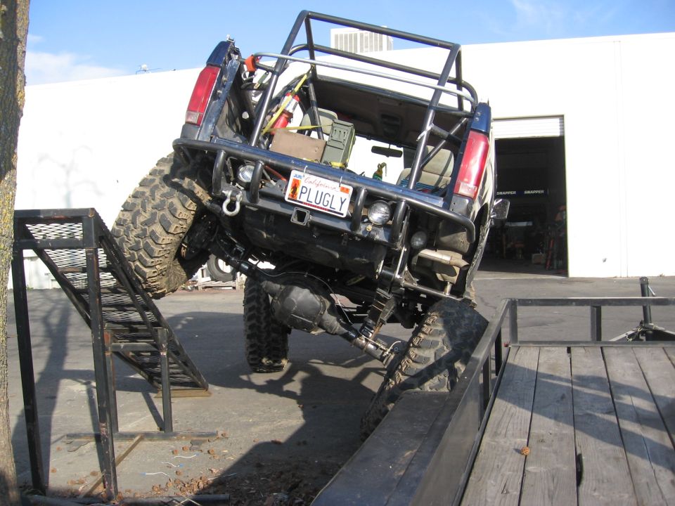 Rear of caged open-top Bronco with PLUGLY plate flexing across ramps outside a shop