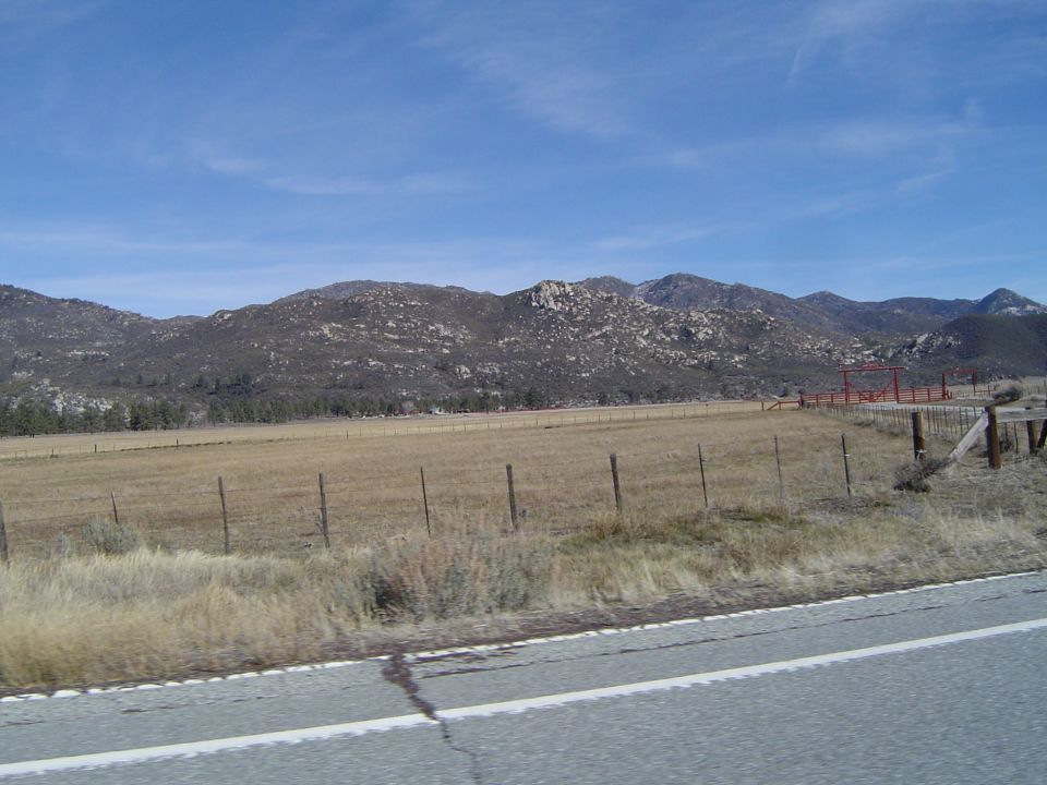 Mountain range under a blue sky with scattered clouds, overlooking a dry grass field with a wooden fence
