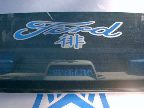 1986 Ford Taurus - A blue Ford emblem is on the rear window above a sticker that reads 'I STOP FOR BLONDES'.