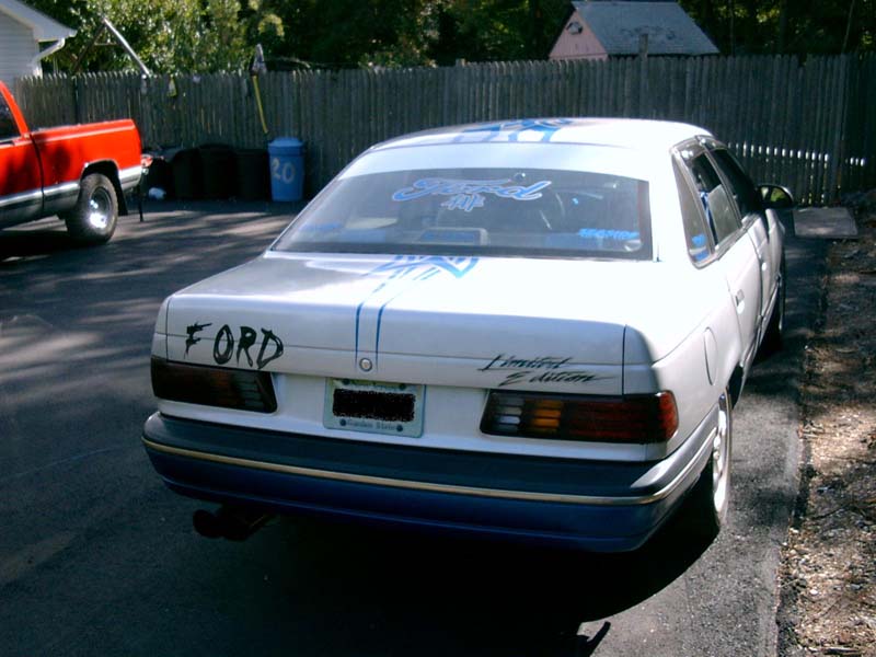 White 1986 Ford Taurus sedan with blue racing stripes and 'FORD' lettering on the trunk