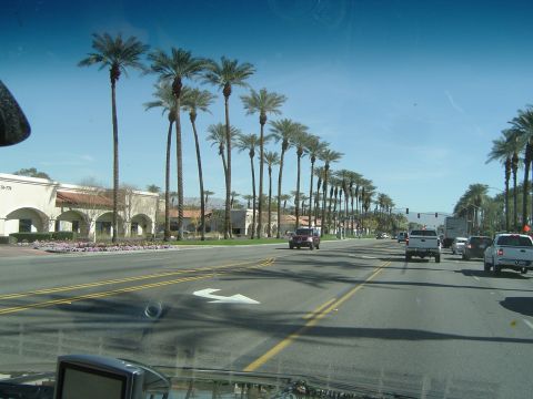 1989 Ford Bronco - A red SUV travels down a street lined with palm trees under a clear blue sky.