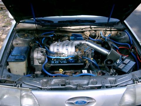 1986 Ford Taurus - The engine bay of a 1986 Ford Taurus features a 3.0 V6 engine with prominent blue hoses and an aftermarket air intake.