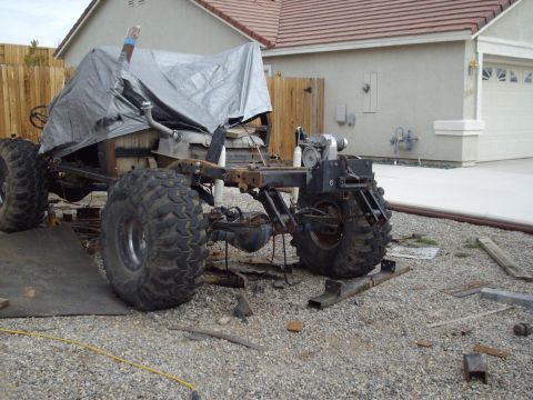 1982 Toyota 4Runner - The chassis and suspension of a vehicle, large off-road tires and a tarp covering the upper portion, sits in a gravel lot.