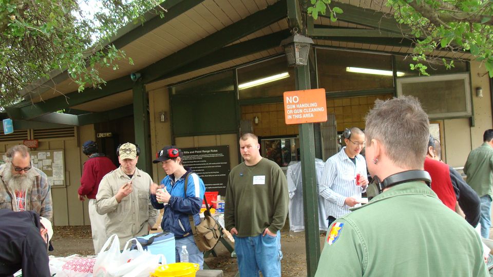 group of people gathered outside a building, some holding items, with trees and a sign that says 'NO GUN HANDLING OR GUN CLEANING IN THIS AREA'