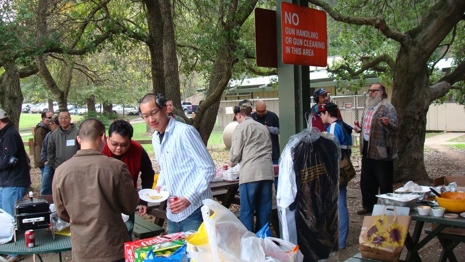 People at picnic tables with food, a grill, and a 'No Gun Handling or Gun Cleaning in This Area' sign