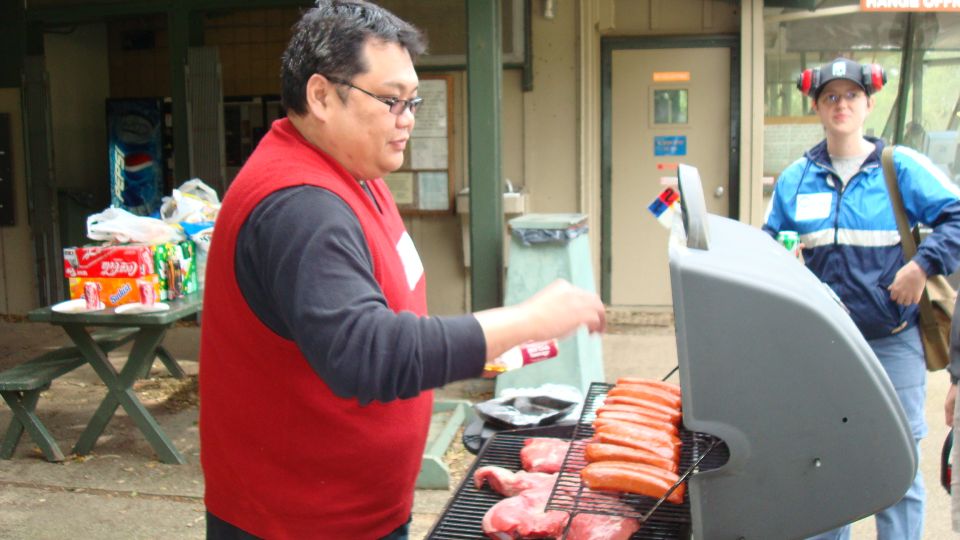 Man in red vest grilling hot dogs and meat on a barbecue with a picnic table and drinks in the background