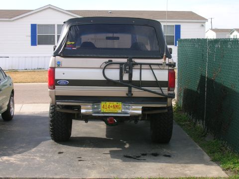 1996 Ford Bronco - Straight-on rear view of a lifted tan Ford SUV with a black soft top, tailgate-mounted swing-out tire carrier (spare removed), chrome rear…