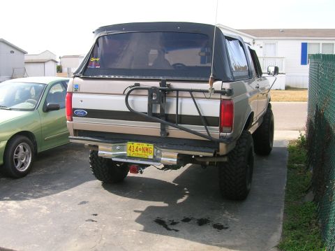 1996 Ford Bronco - The rear of a tan and silver Ford Bronco with a black soft top and a spare tire carrier is visible.