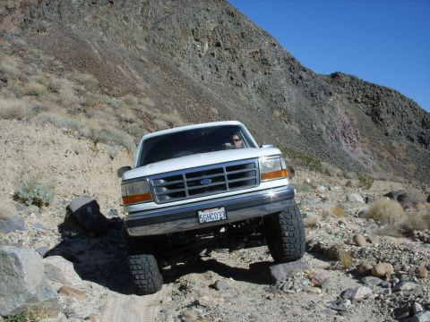 1993 Ford Bronco - A white Ford truck is shown driving over rocks on a steep, arid mountainside under a clear blue sky.