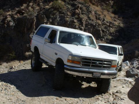 1993 Ford Bronco - Two white SUVs navigate a rocky, off-road path under a bright sun.