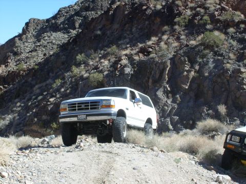 1993 Ford Bronco - A white Ford Bronco navigates a rough, mountainous terrain, with another vehicle partially visible on the right.