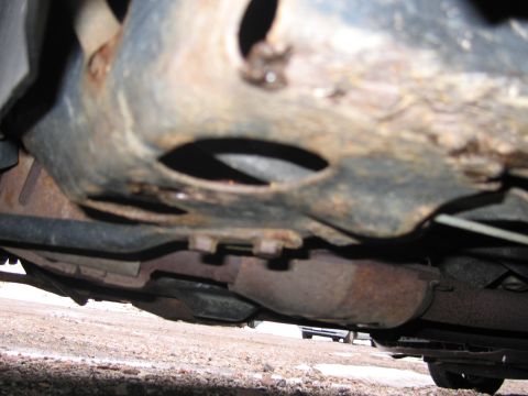 2001 Ford Crown Victoria - Close-up view of the underbody of a vehicle, revealing rusted frame components and exhaust system parts. A second vehicle is faintly visibl…