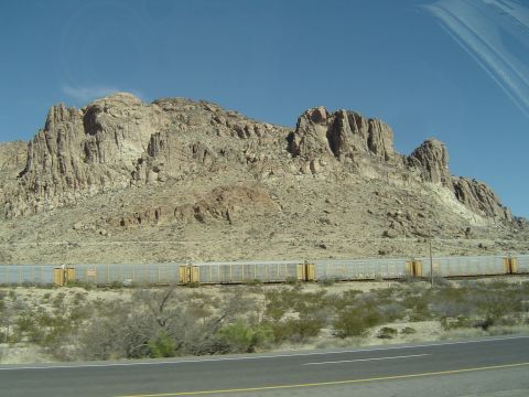 1989 Ford Bronco - A long train travels through a vast desert landscape, dwarfed by mountains under a bright blue sky.