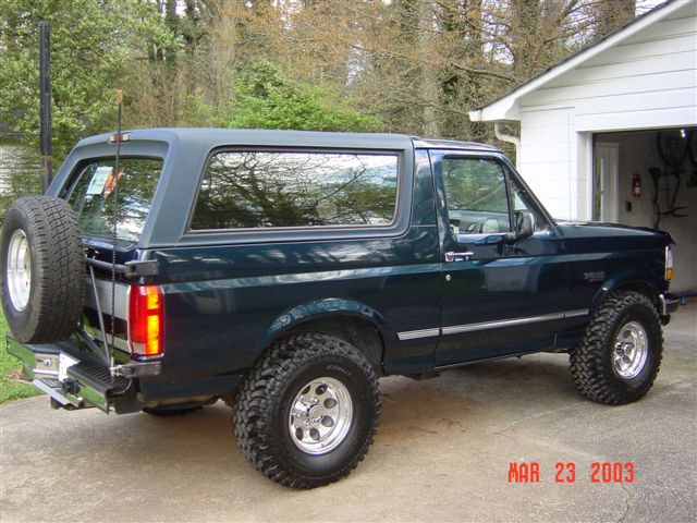 Dark green Ford Bronco SUV with rear spare tire and off-road tires parked by a white garage