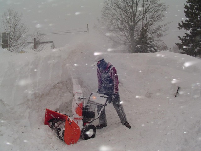 Orange snow blower clearing snow with a person operating it during a snowstorm