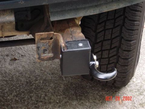 1995 Ford Bronco - Underbody close-up of a fabricated black steel box bracket with a bolt-on tow hook welded to the rusty frame rail, just ahead of an all-ter…